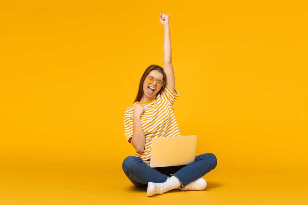Joyful girl with laptop computer, sitting on a floor and celebrating something that happenend on-screen, isolated on yellow background