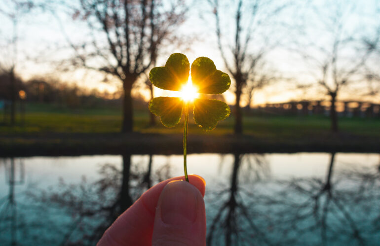 Hand holding a single four leaf clover in front of sunrise on lake