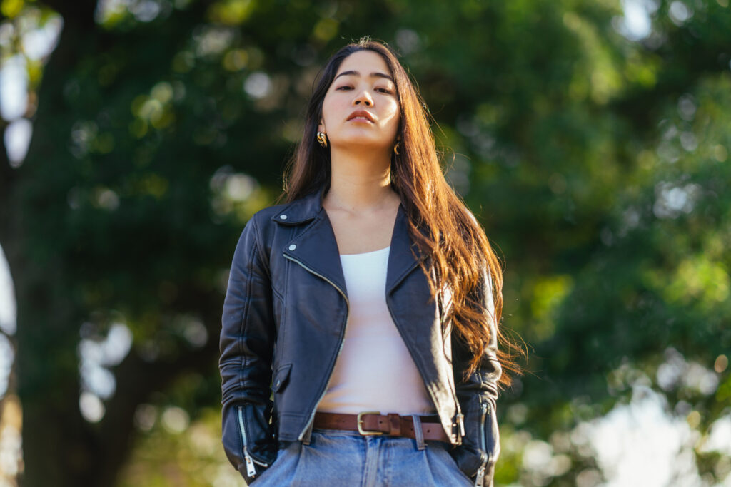 Young, confident woman in leather jacket standing outside with hands in pockets looking directly at camera